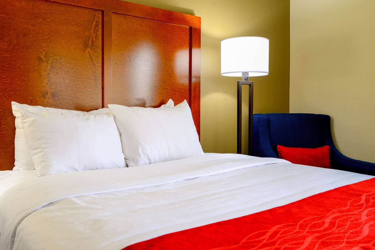 Habitación de hotel con una cama grande, cabecera de madera, almohada decorativa roja y una silla azul, además de una lámpara de pie.