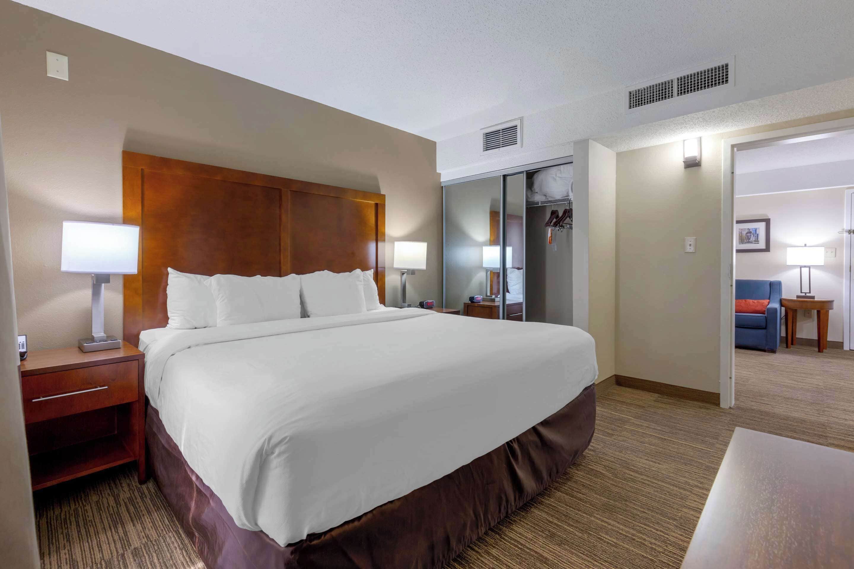 A hotel room with a large bed, a nightstand, a lamp, a wardrobe, and a desk with a chair.
