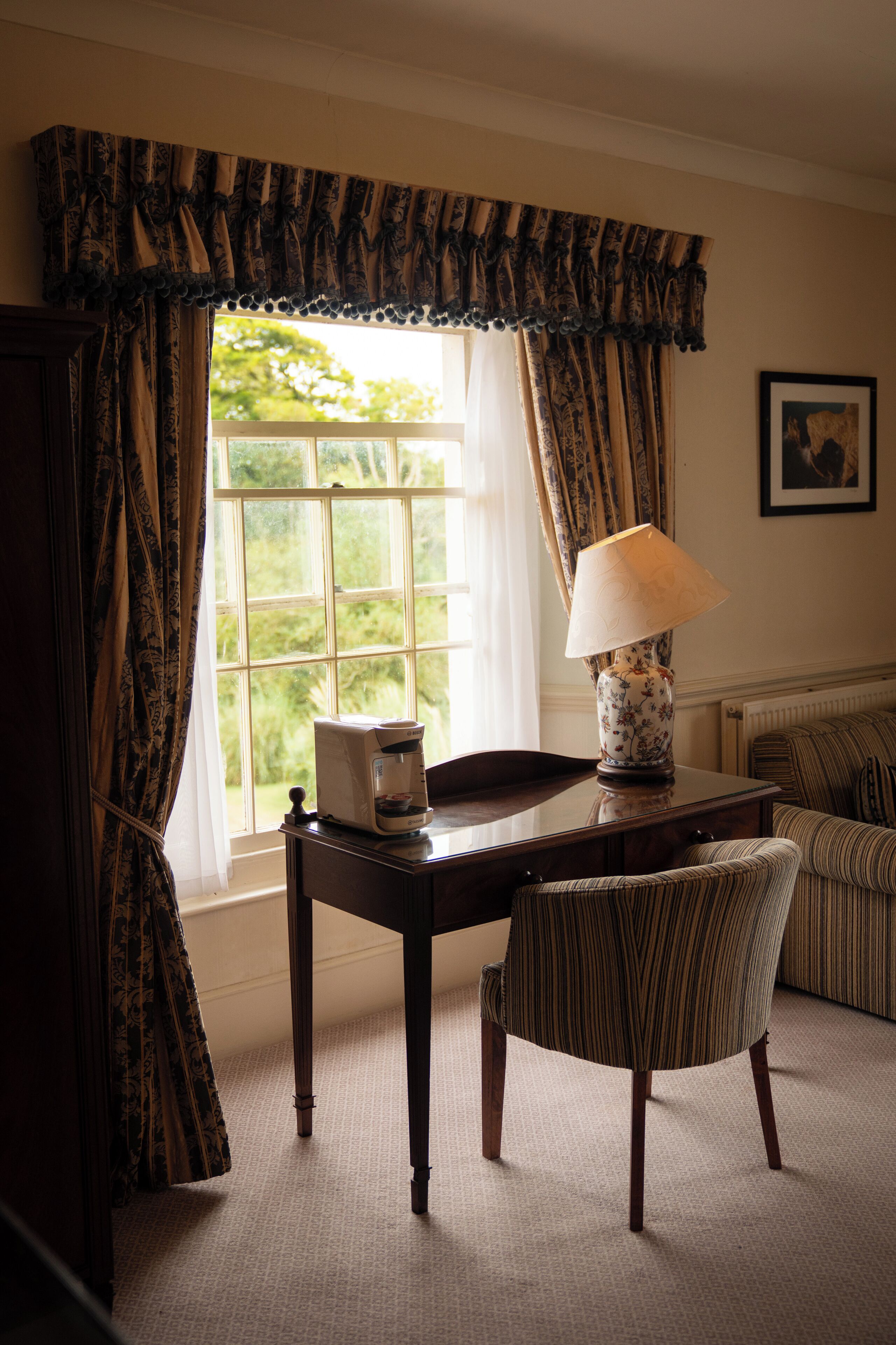 A room with a window, a table with a lamp, a chair, and a framed picture on the wall.