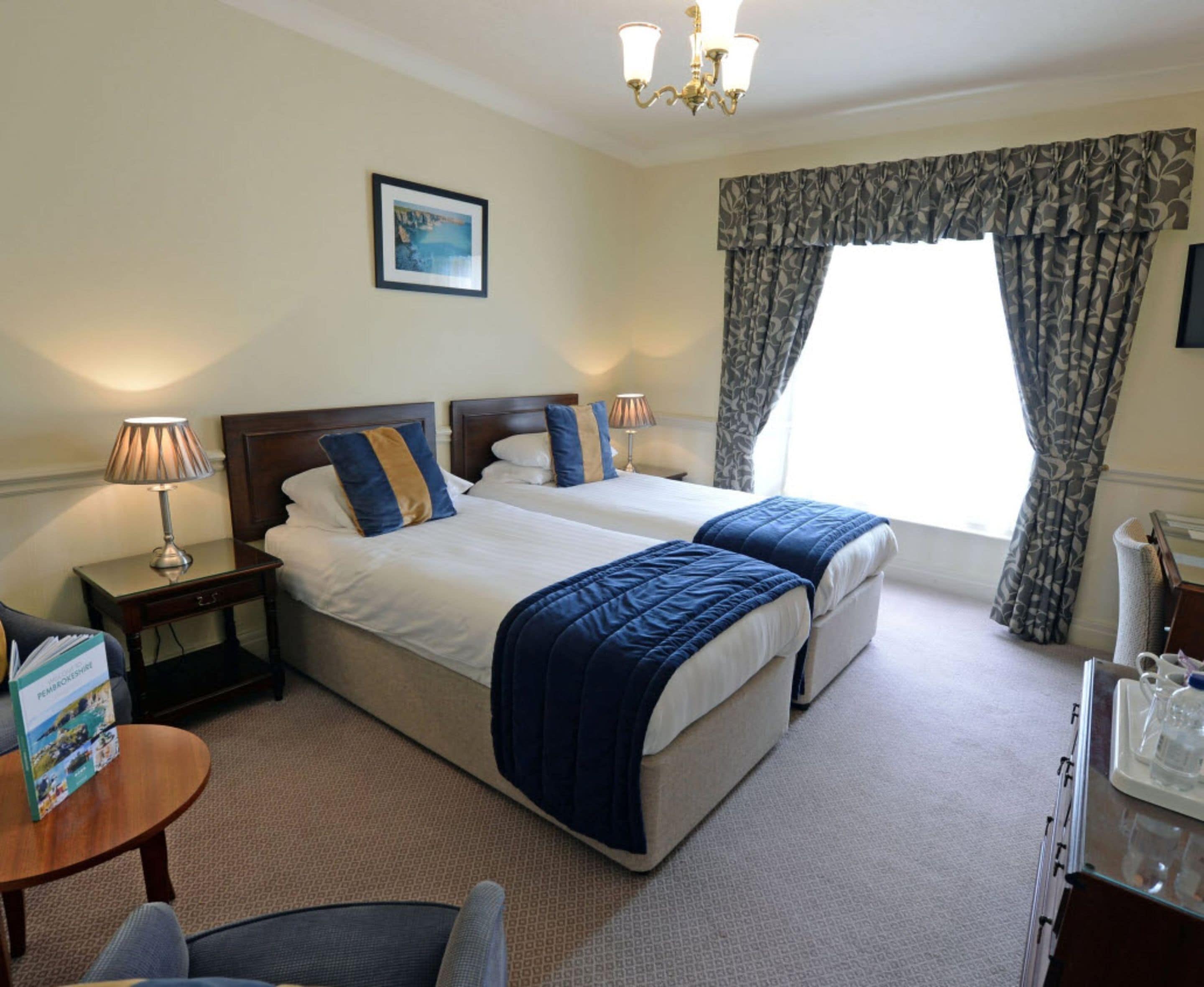 A hotel room with two beds, a bedside table with a lamp, a small round table with a book, and a window with curtains.