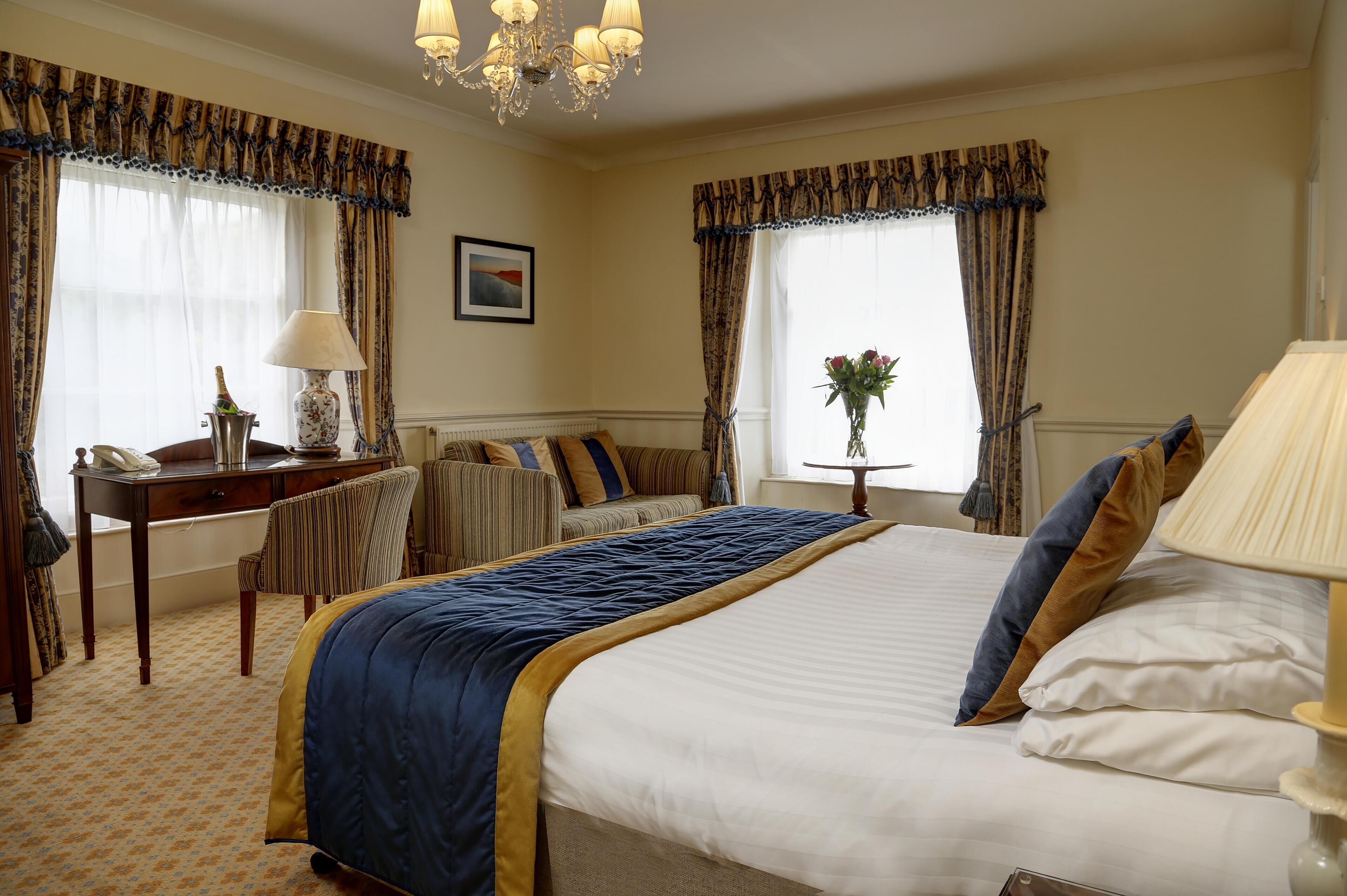 A hotel room with a large bed, a desk with a lamp, a chair, and a small table with a vase of flowers.