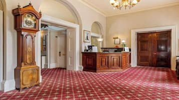 A grand hallway with a large wooden clock, a reception desk, and a red patterned carpet.