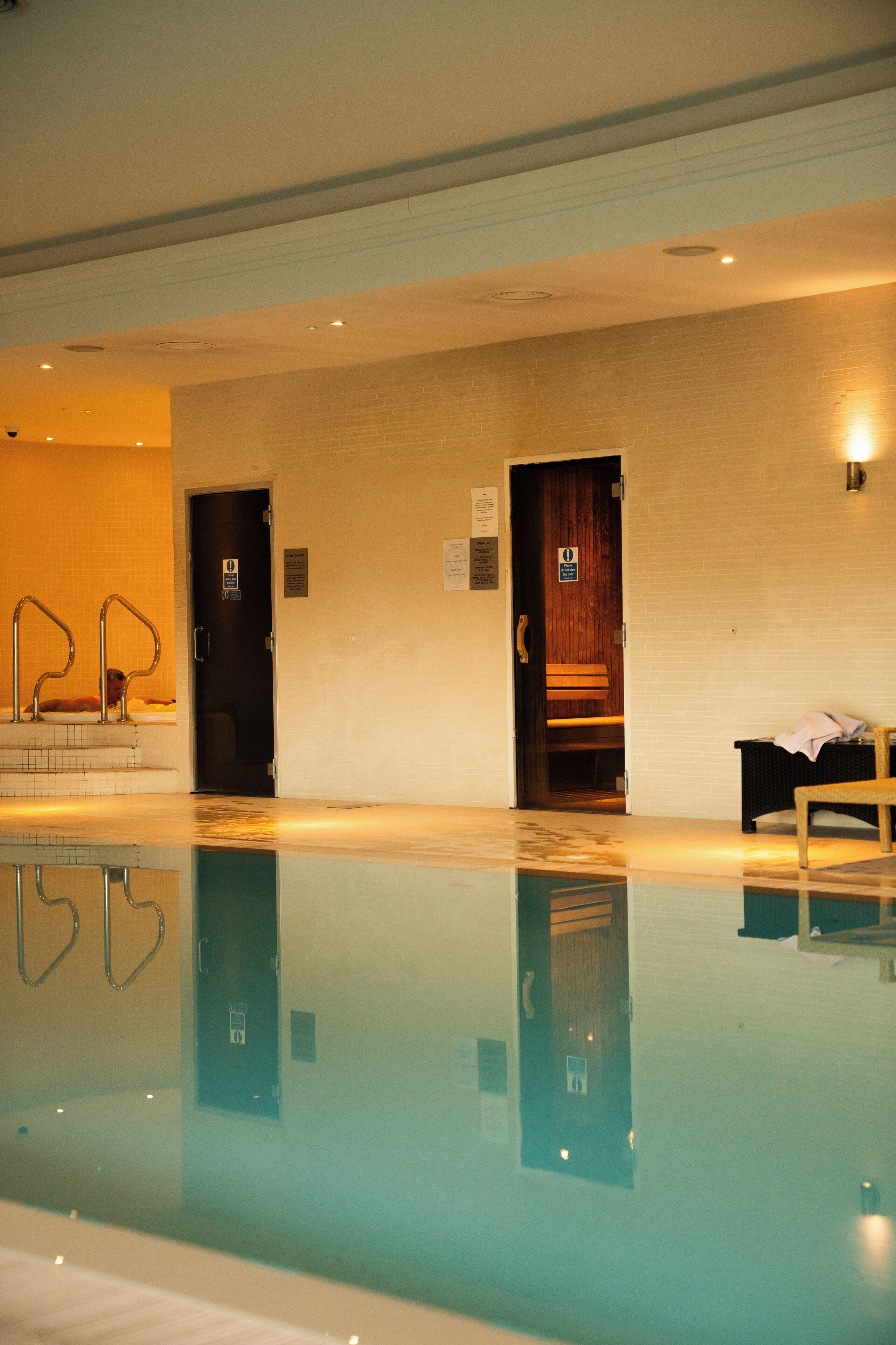 A swimming pool with a clear reflection, a wooden bench, and a door leading to a changing room.