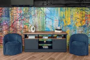 A room with a large abstract painting, a wooden table, two blue armchairs, and a dark-colored cabinet with books. - ARC Hotel, Washington DC, Series by Marriott (Washington)