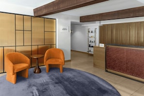 A reception area with a wooden reception desk, two orange chairs, a small round table, and a blue carpet. - ARC Hotel, Washington DC, Series by Marriott (Washington)