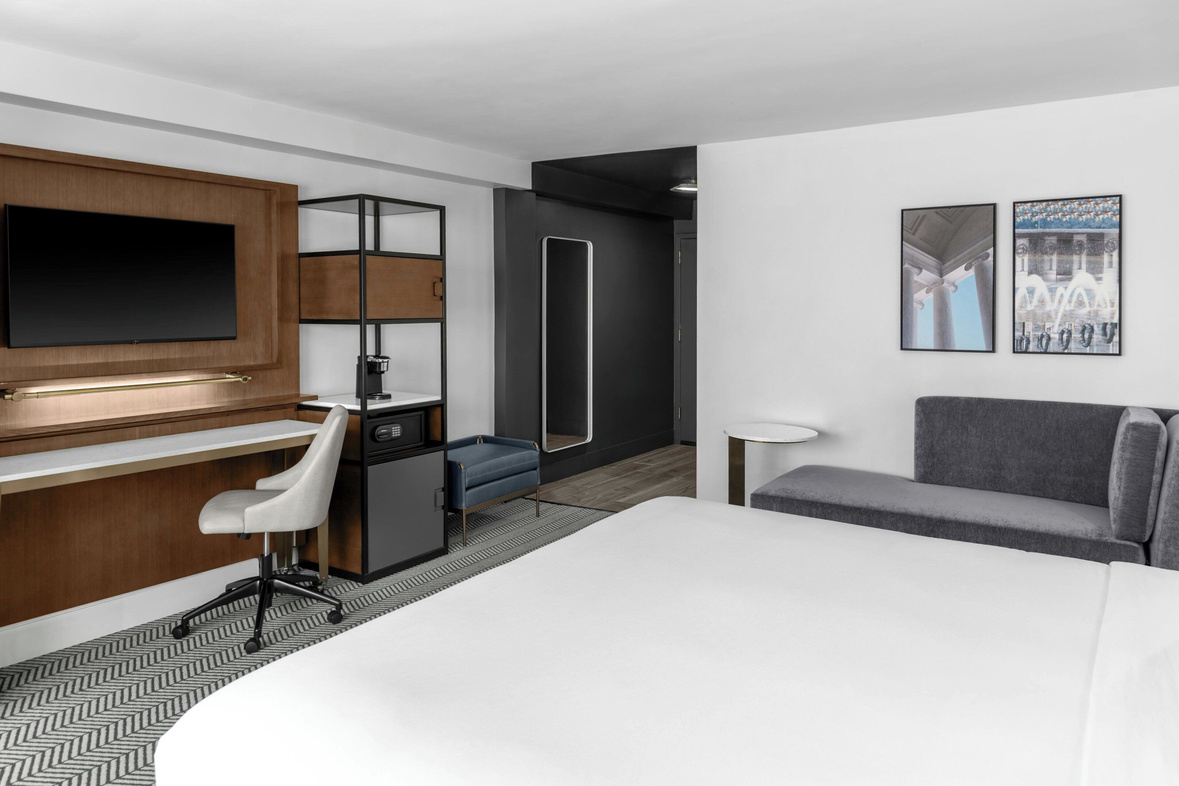 A modern hotel room with a large bed, a desk with a chair, a small sofa, and a TV mounted on the wall.