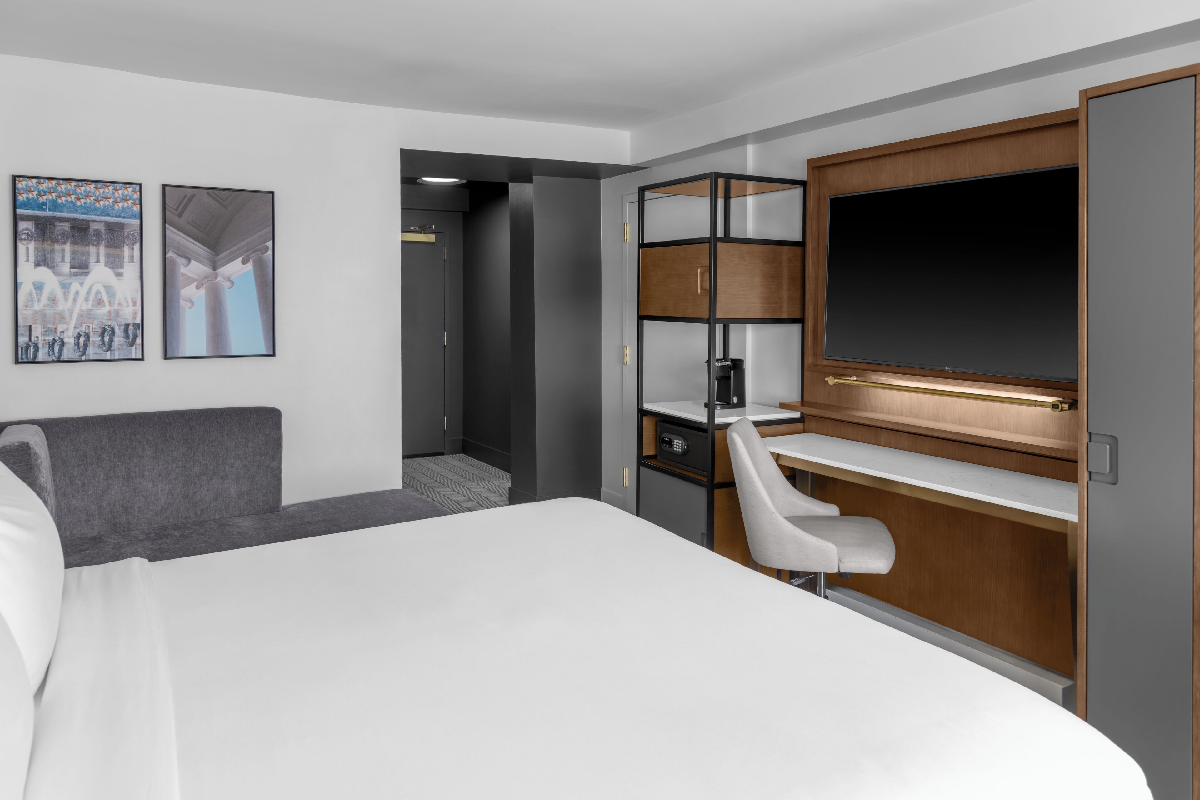 A hotel room with a bed, a desk, a chair, a TV, and a door leading to a hallway.