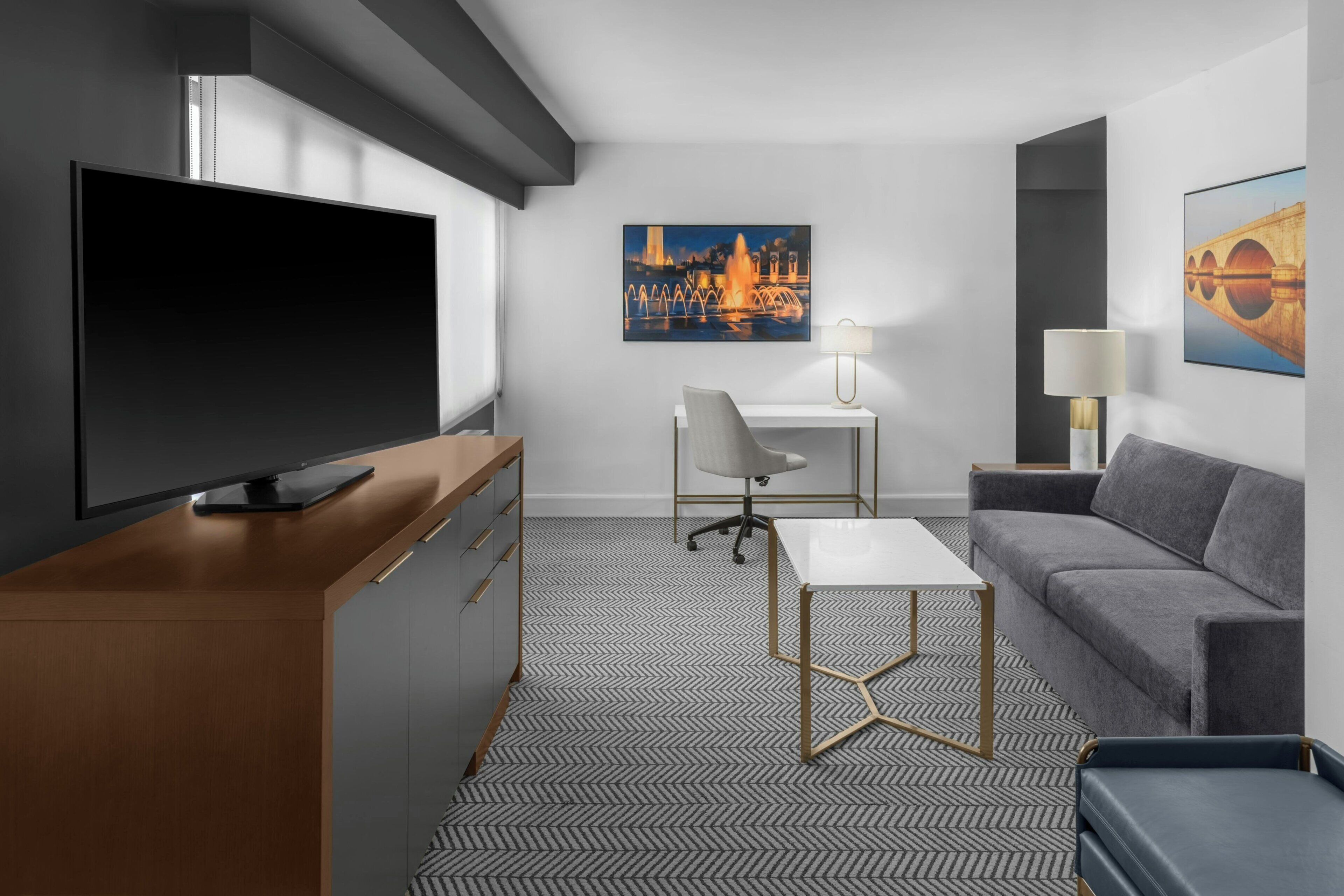 A modern hotel room with a flat-screen TV, a desk, a sofa, and two paintings on the walls.