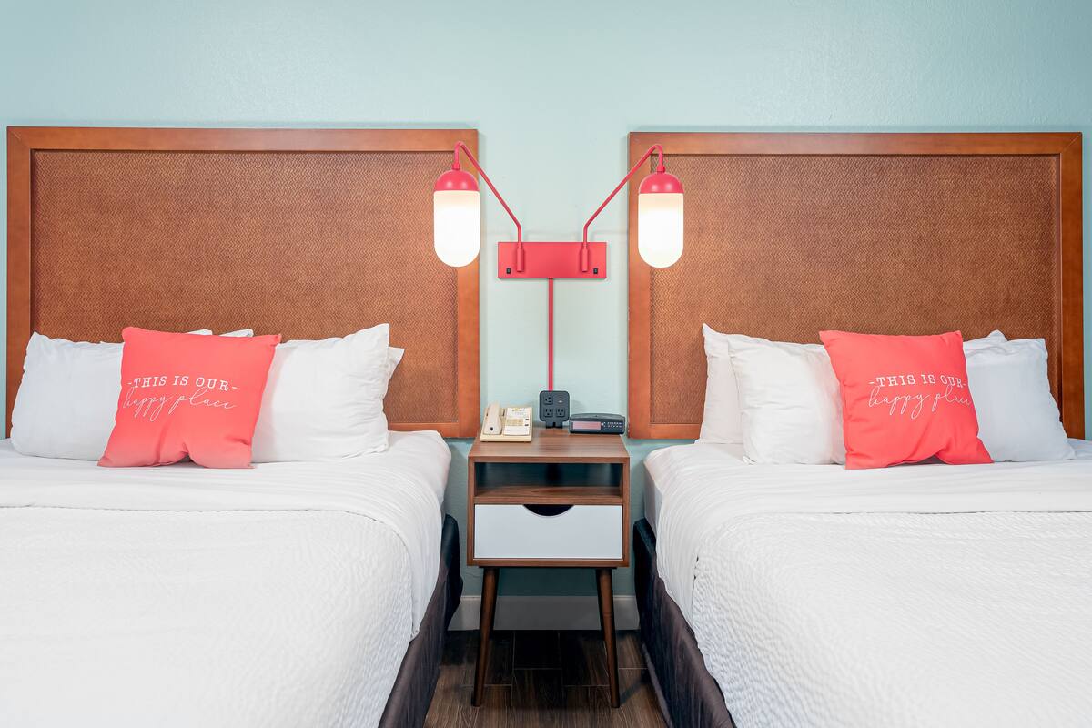 A hotel room with two beds, each with white bedding and a pink pillow. There is a nightstand between the beds with a lamp and a phone.