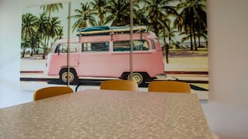 Un comedor con una mesa y sillas, una furgoneta rosa con tablas de surf al fondo y palmeras.