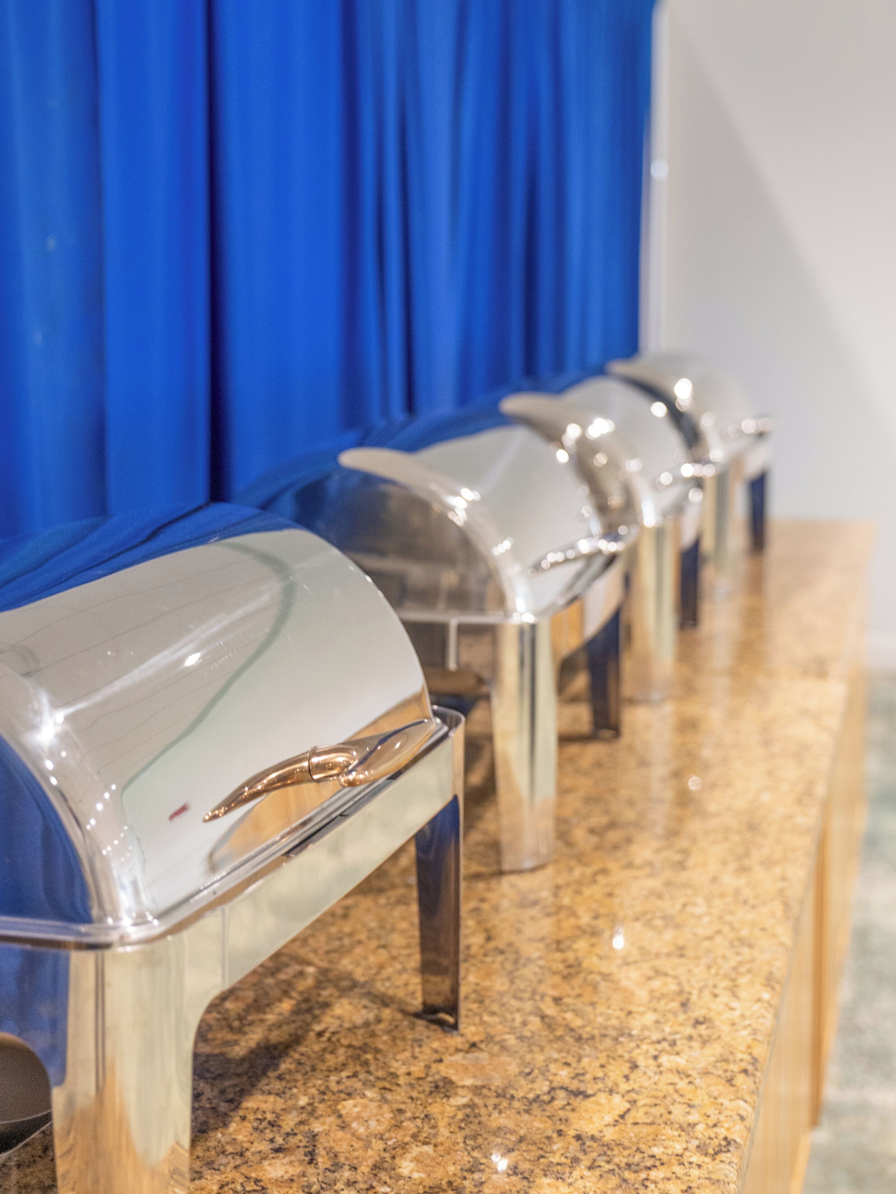 Uma fileira de estações de buffet com tampas de vidro transparente e puxadores de aço inoxidável em bancada de granito.