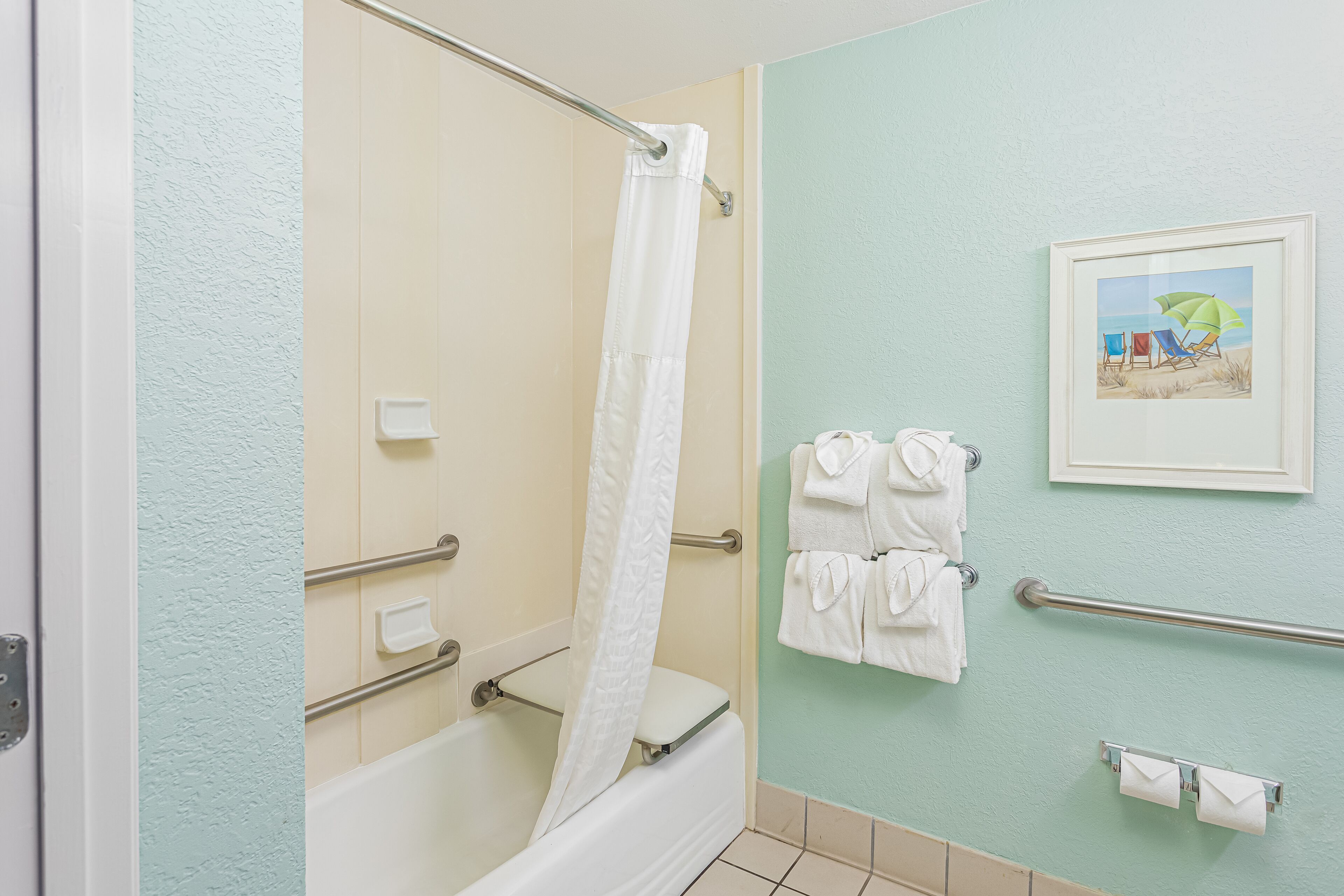 A bathroom with a bathtub, a shower curtain, a towel rack with towels, a framed picture on the wall, and a handrail.
