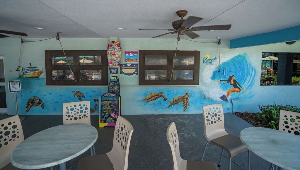 A restaurant with a mural of a woman surfing and sea turtles, tables, chairs, and a ceiling fan.