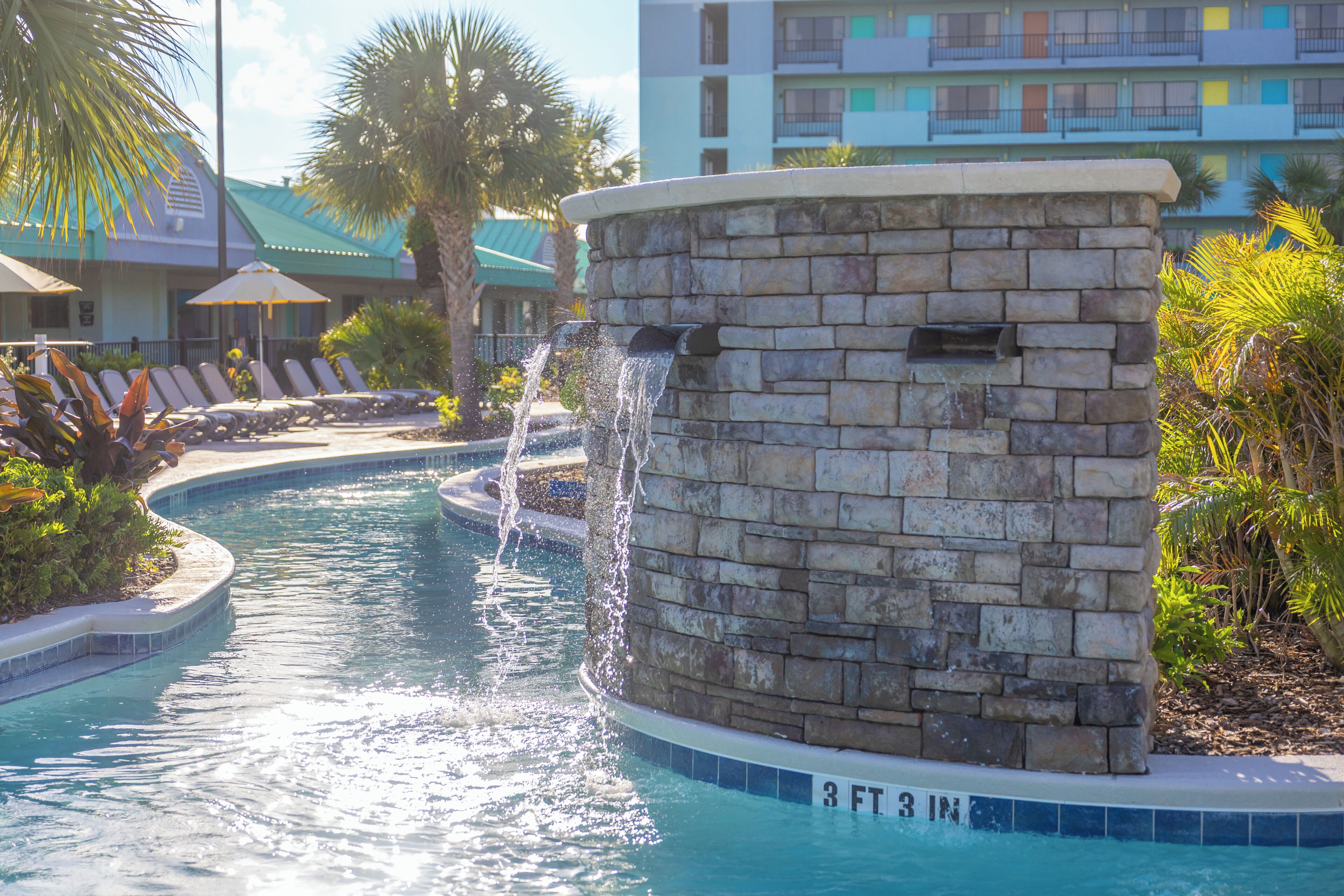 A swimming pool with a stone wall, lounge chairs, and a building with balconies in the background.