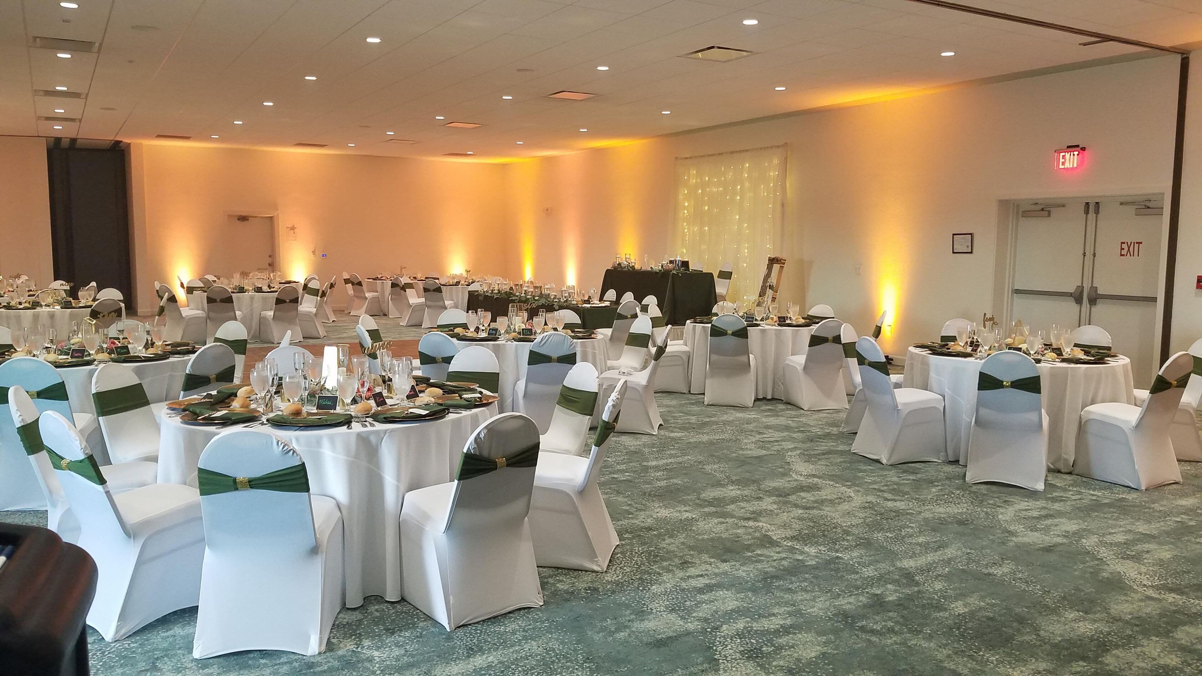 Een balzaal met ronde tafels voor een formeel evenement, witte stoelen met groene linten, en een vloerbedekking.