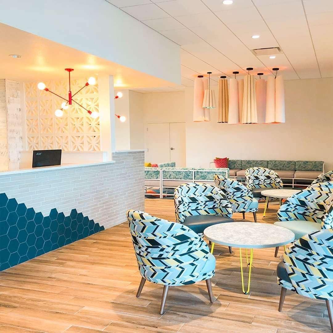 Un área de lounge moderna con paredes de diseño hexagonal, un mostrador de recepción y una zona de estar con sillas estampadas y mesas redondas.