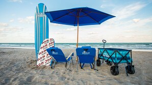 A beach scene with a blue umbrella, two beach chairs, a surfboard, and a beach cart. - Beachside Hotel & Suites Cocoa Beach - Port Canaveral (Cocoa Beach)