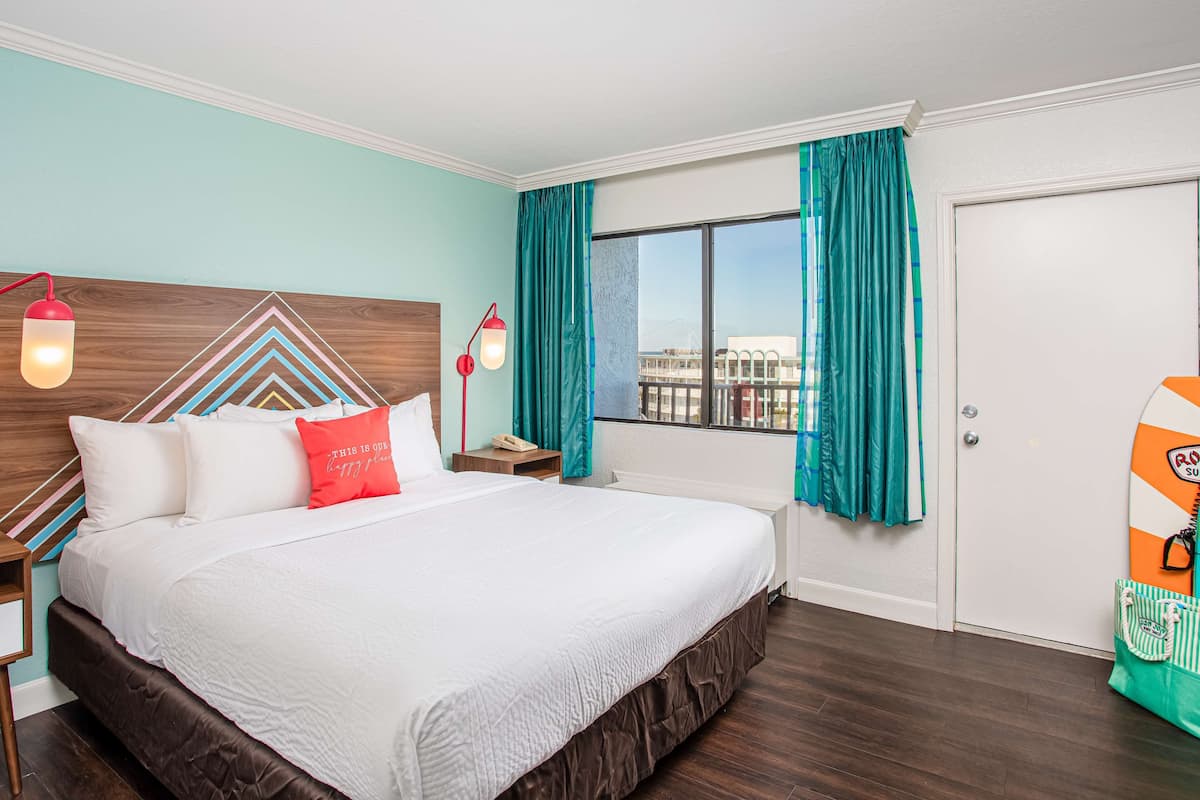 A hotel room with a large bed, a wooden headboard, teal curtains, a window with a view, and a surfboard on the wall.