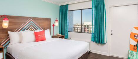 Une chambre d’hôtel avec un grand lit, une tête de lit en bois, des rideaux turquoise, une fenêtre donnant sur un beau paysage et une planche de surf accrochée au mur.