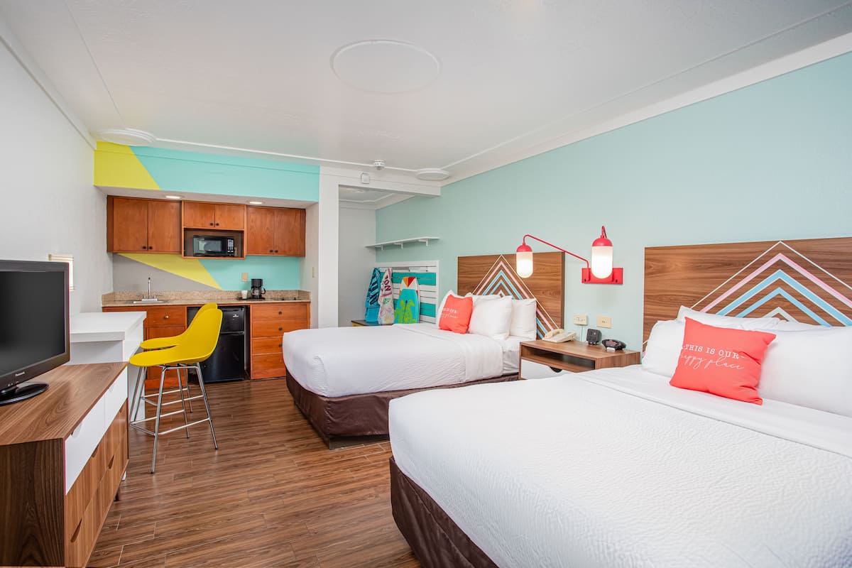 A hotel room with two beds, a kitchenette, a TV, and a bright yellow chair.