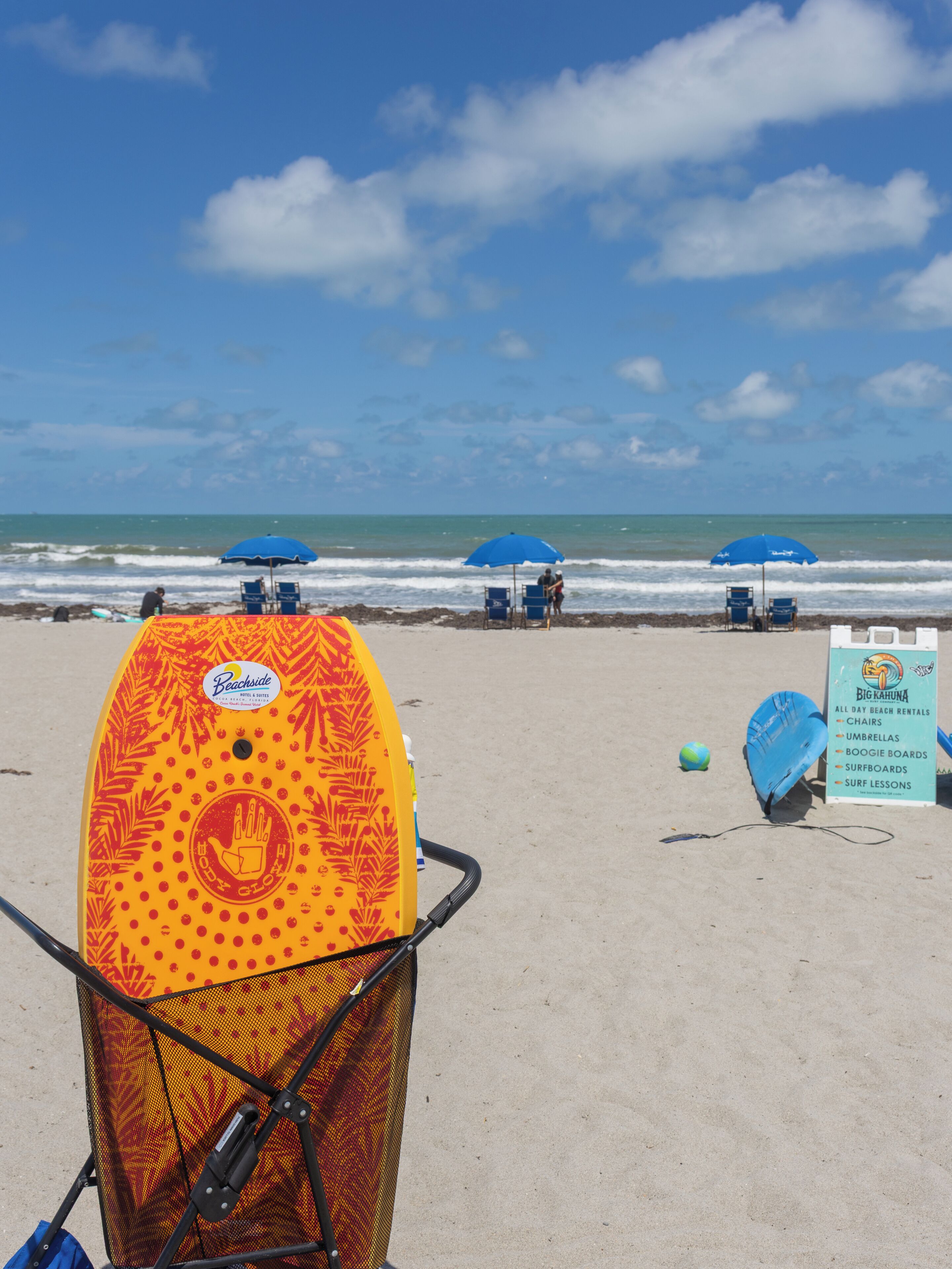 Una spiaggia con ombrelloni, un cartello per il noleggio di attrezzature da spiaggia e una tavola da surf.