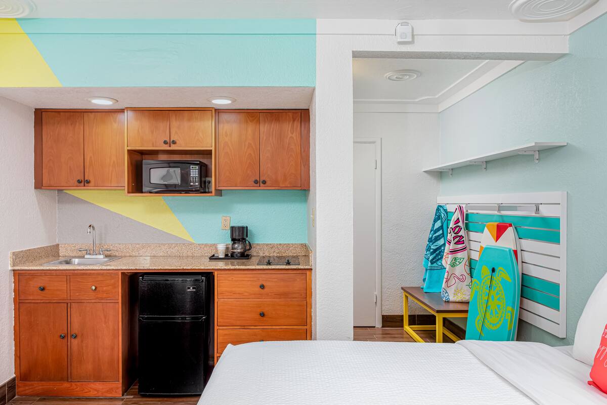 A compact hotel room with a kitchenette, a bed, and surfboards against the wall.