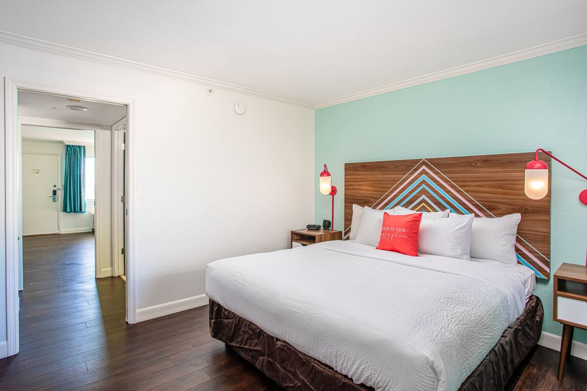 A neatly made bed with a wooden headboard, white bedding, and a red pillow. There are two bedside tables with lamps. The room has a wooden floor and a teal accent wall.
