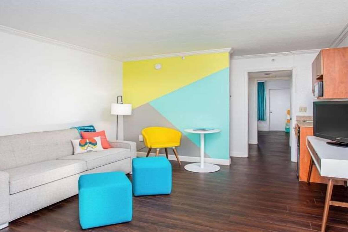 A modern living room with a grey sofa, two blue ottomans, a yellow chair, a small white table, and a TV on a wooden stand.