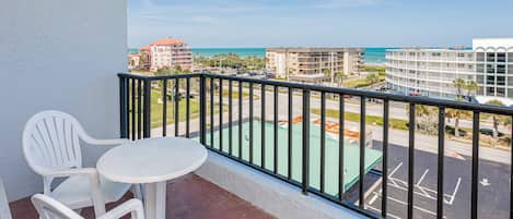 A balcony with a white table and two chairs, overlooking a parking lot and buildings with a view of the ocean.