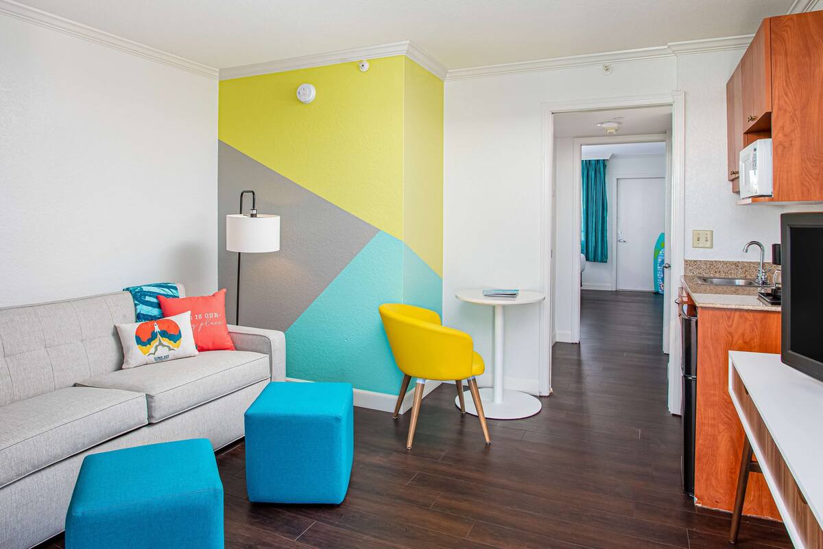 A modern living room with a sofa, colorful cushions, a bright wall, and a kitchen area in the background.