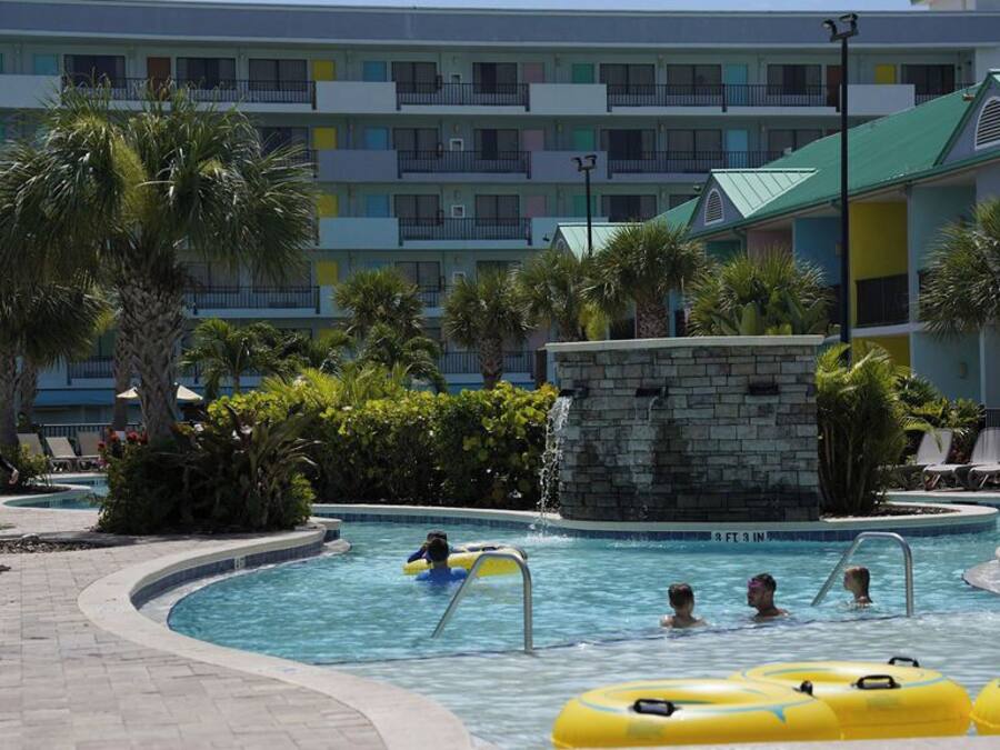 Zona de piscina de hotel con varios niveles de balcones, palmeras y sillones de descanso.