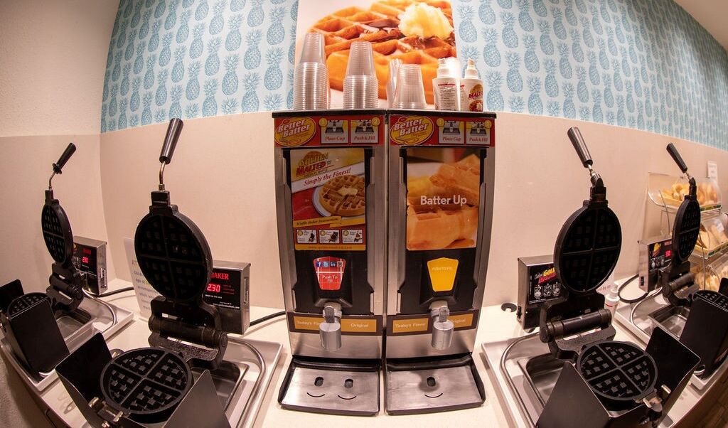 A waffle station with multiple waffle makers, a display of waffles, and a sign for 'Batter Up'.