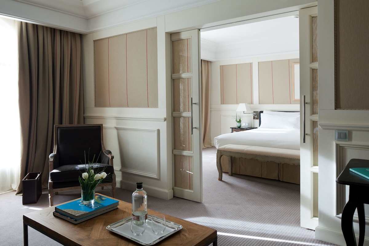 Une chambre d’hôtel avec un lit, une chaise, une table avec un livre et un vase, et une table de chevet avec une lampe.