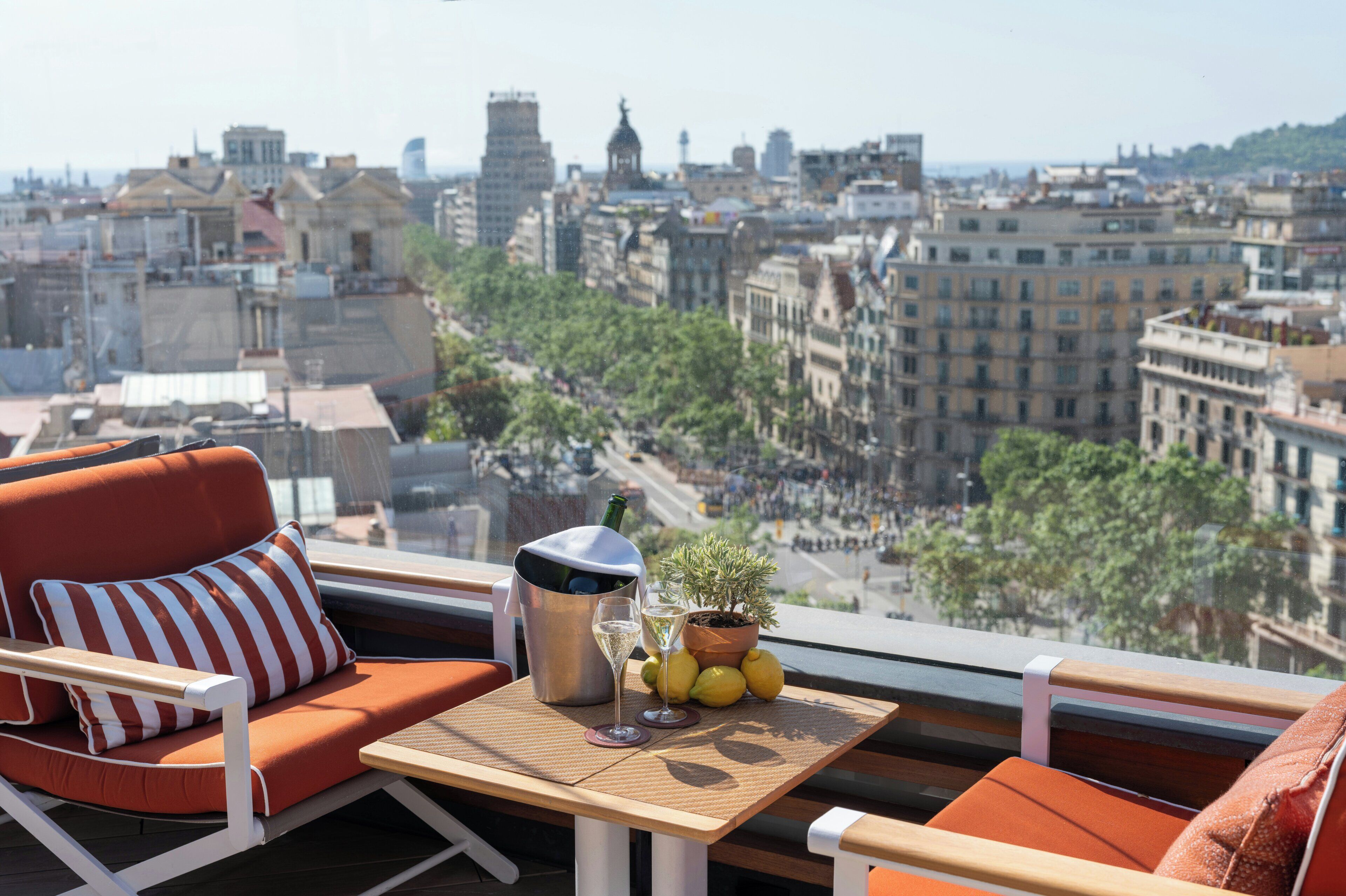 A balcony with a view of a cityscape, a table with a bottle, glasses, and a plant.