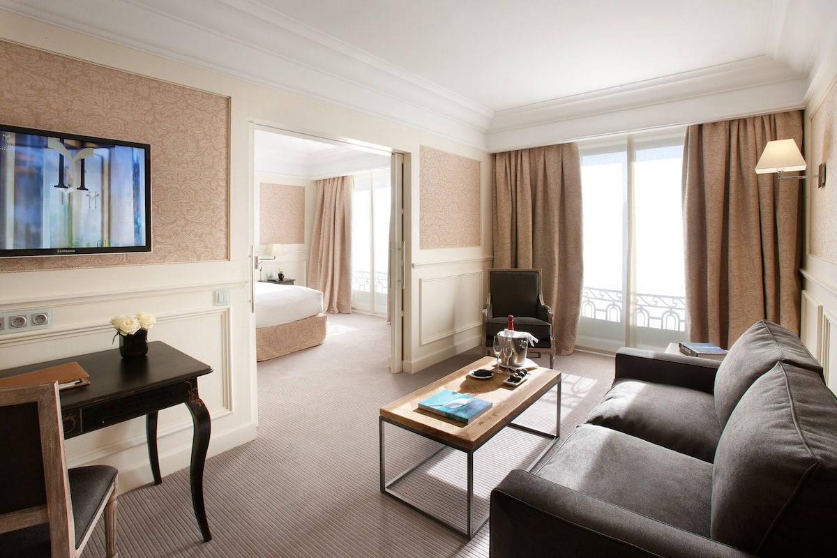 A hotel room with a flat-screen TV, a wooden coffee table, a grey sofa, a desk with a chair, and a bed in the background.