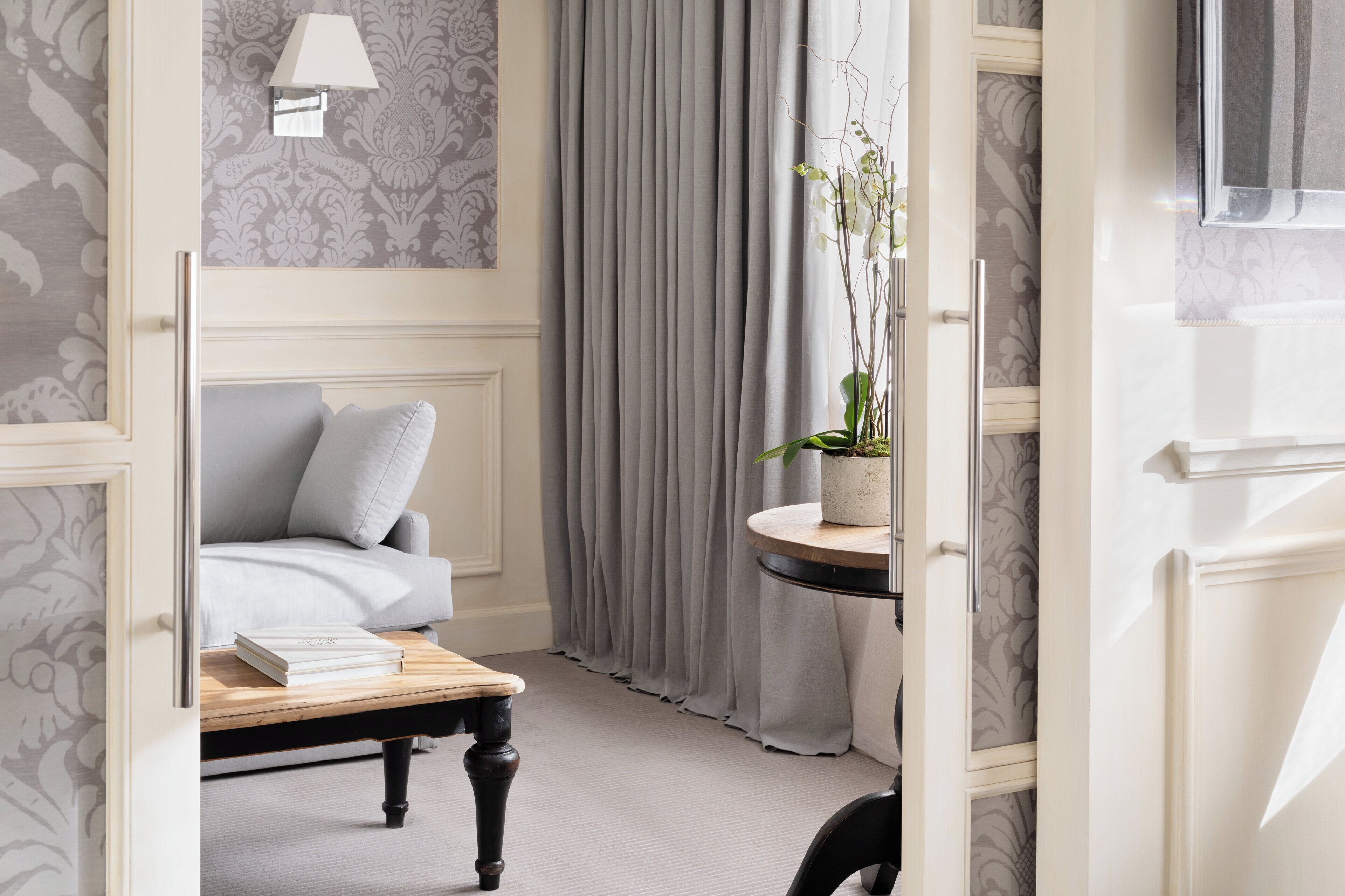Una habitación con una cama, una mesita de noche con libros, una planta en maceta y una ventana con cortinas.