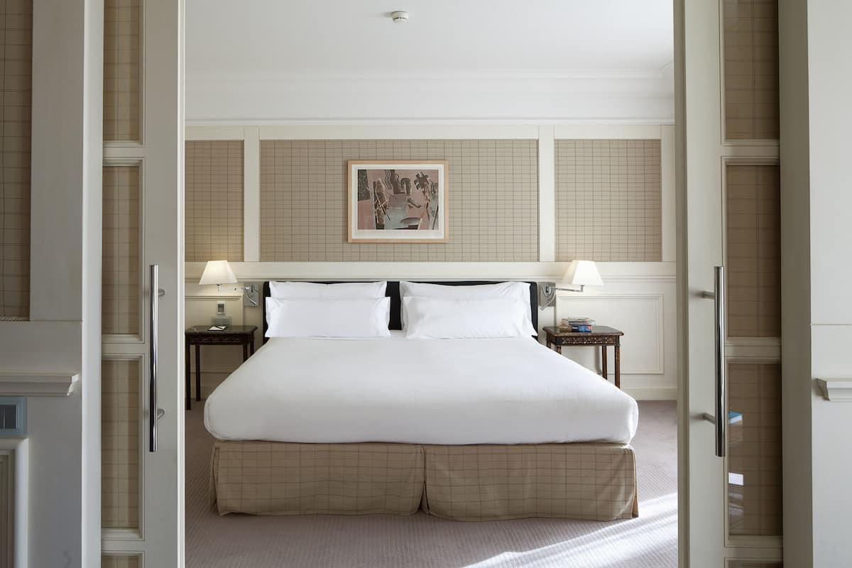 A hotel room with a large bed, two bedside tables with lamps, a framed artwork on the wall, and a checkered floor runner.