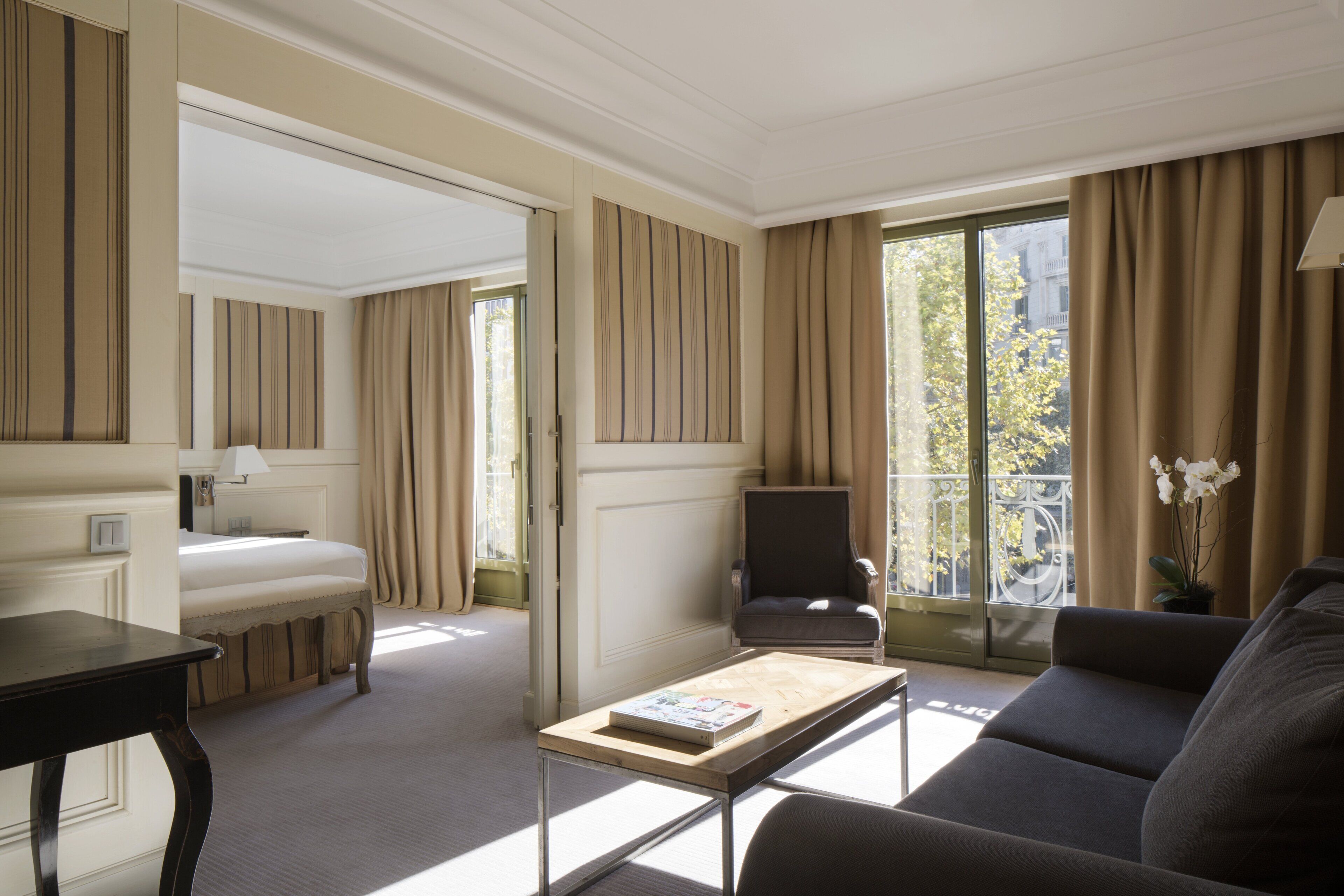 Habitación de hotel con cama, sofá, mesa de centro, sillón y un amplio ventanal con cortinas.