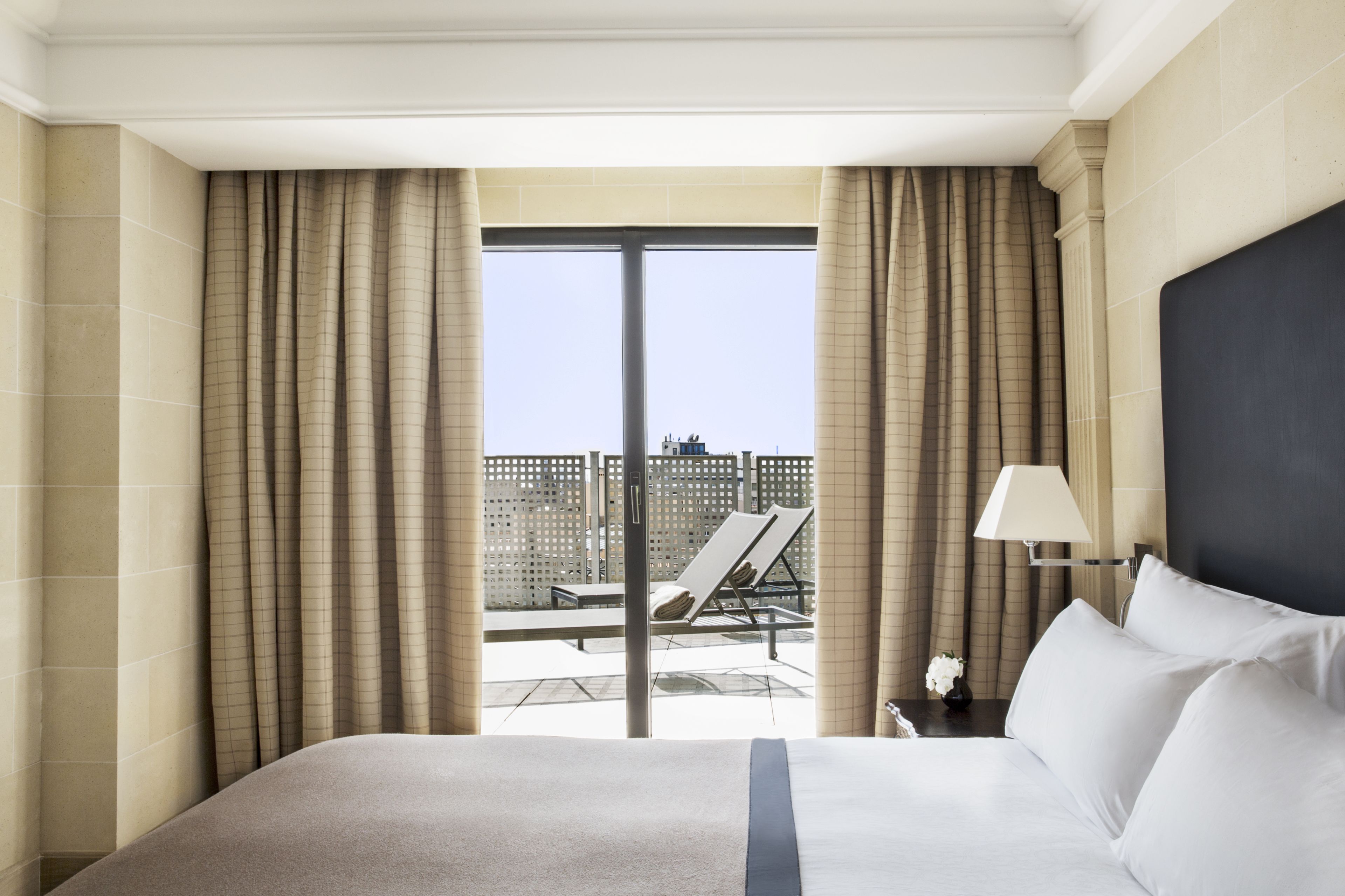 A hotel room with a large bed, a view of the city through floor-to-ceiling windows, and a balcony with a chair and table.
