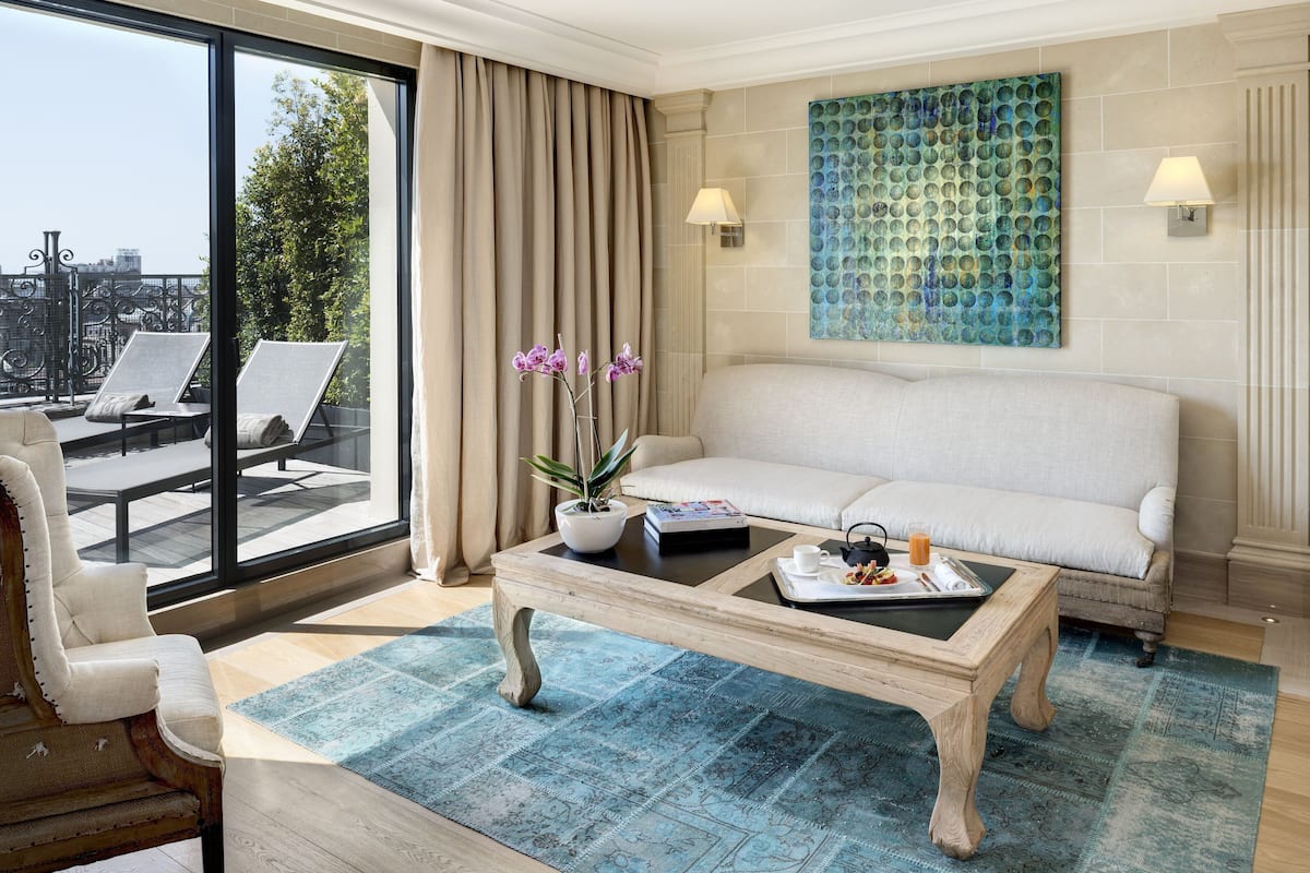 A hotel room with a large window, a sofa, a coffee table with a tray, a chair, and a decorative wall piece.
