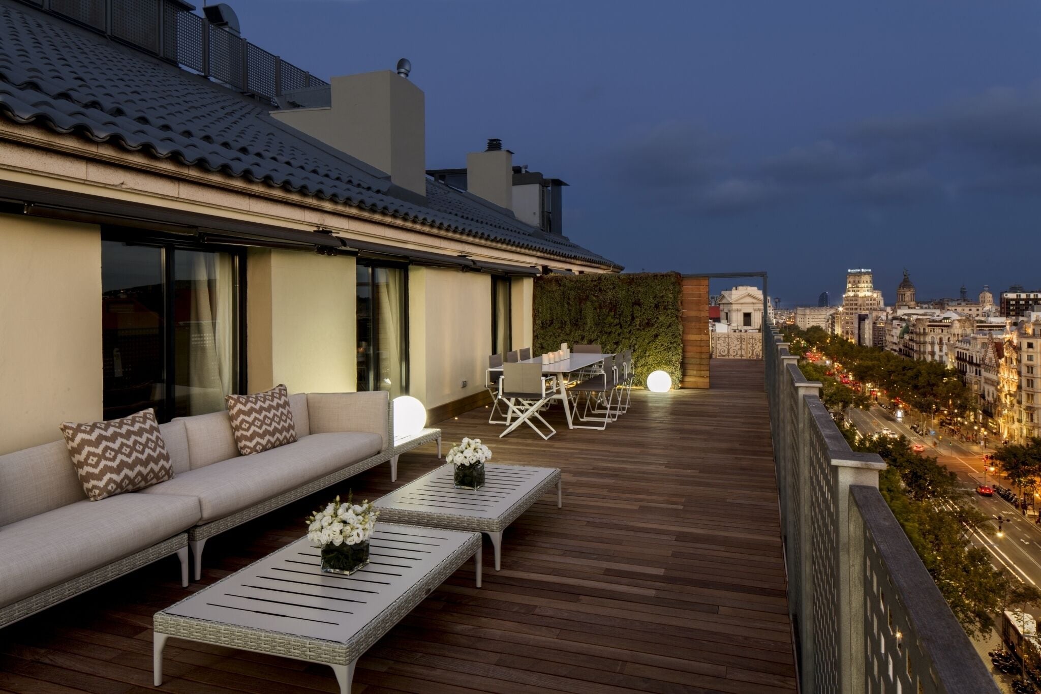 A rooftop terrace with a sofa, dining table, and chairs, overlooking a cityscape at dusk.