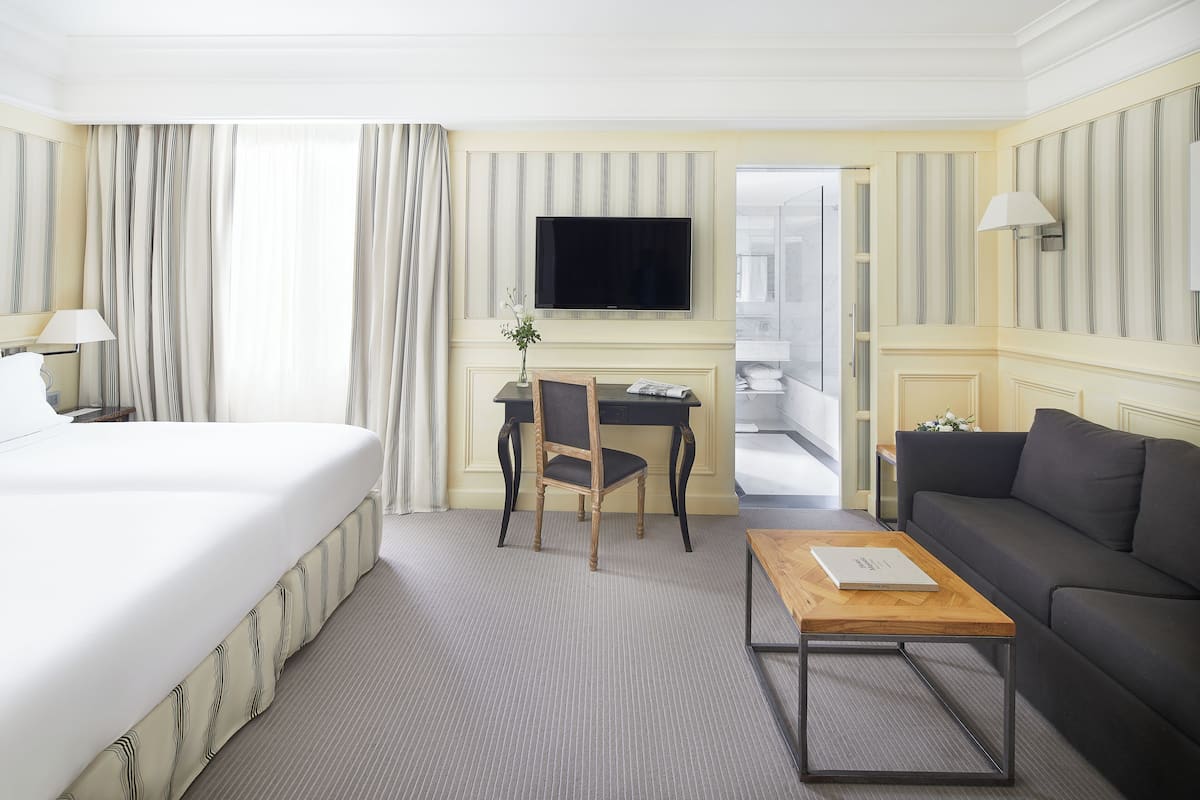 A hotel room with a bed, a desk, a chair, a sofa, a TV, and a lamp.