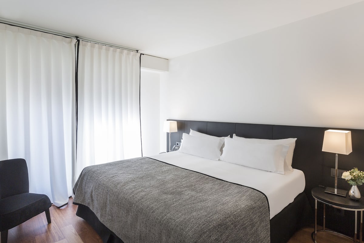 A hotel room with a bed, a chair, a bedside table with a lamp, and a window with curtains.