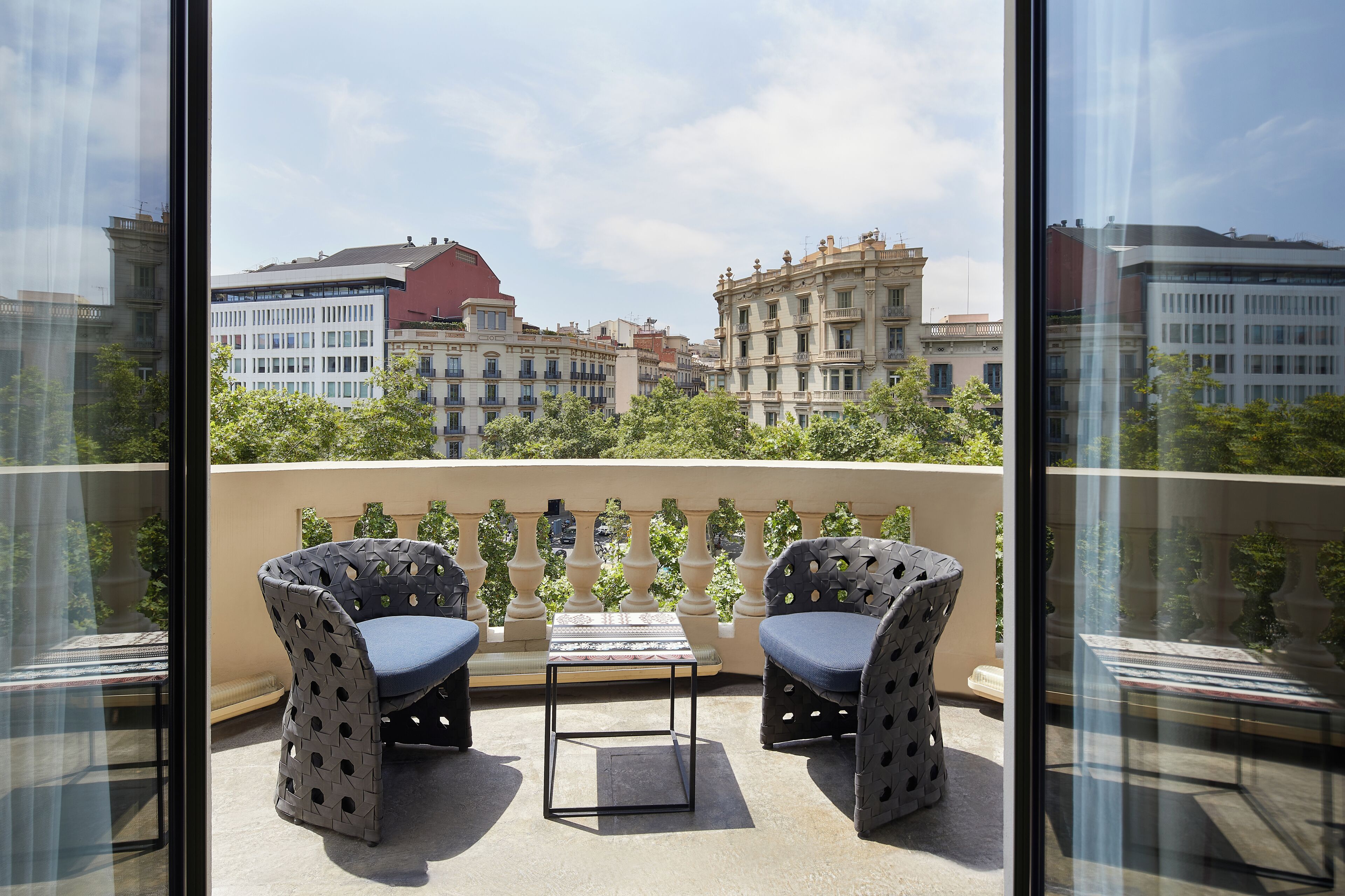 A balcony with two patterned chairs and a small table, overlooking a cityscape with historic buildings.