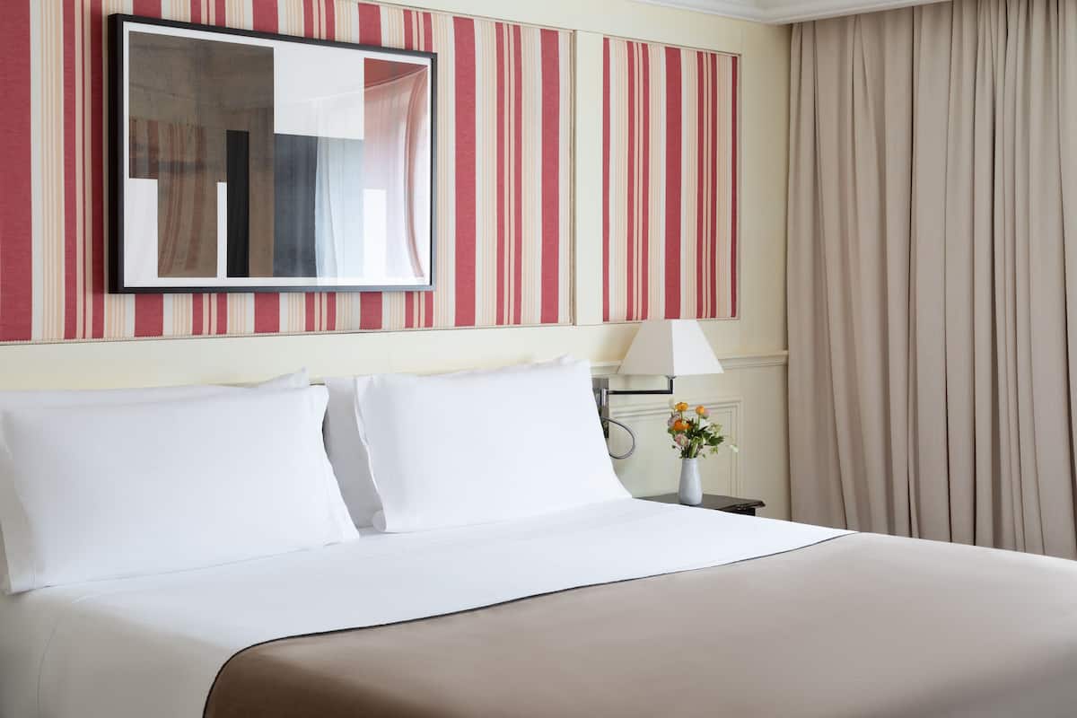 A hotel room with a bed, a nightstand, a lamp, a vase with flowers, and a striped wall.