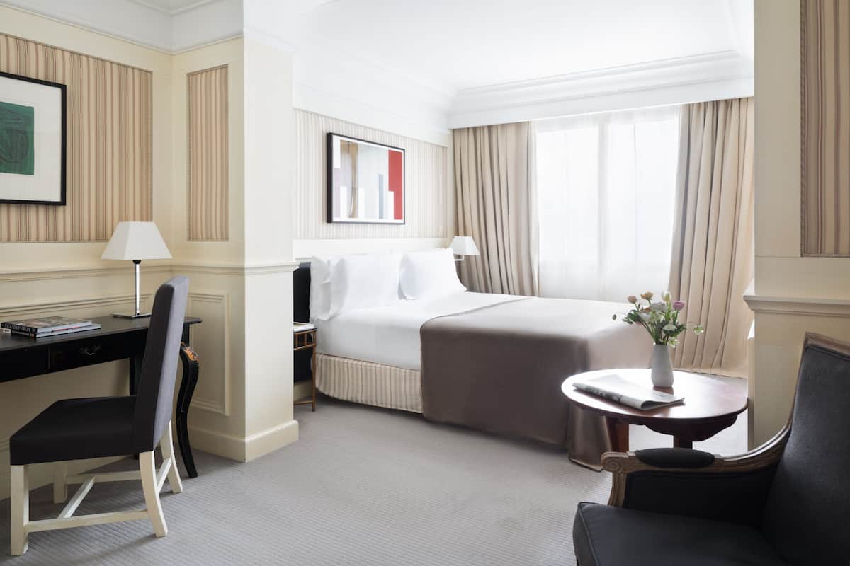 A hotel room with a bed, a desk with a chair, a small table with a vase, and a window with curtains.