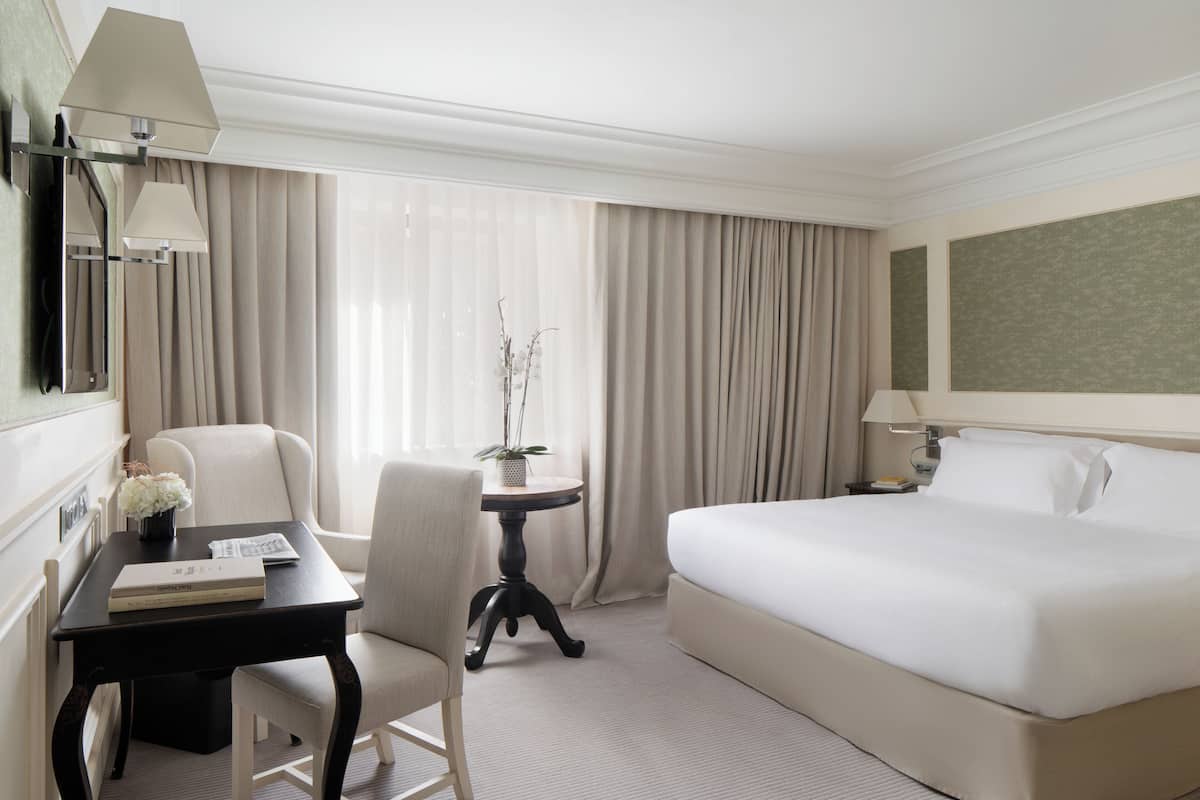 A hotel room with a bed, a desk with a chair, a small table with a plant, and a window with curtains.