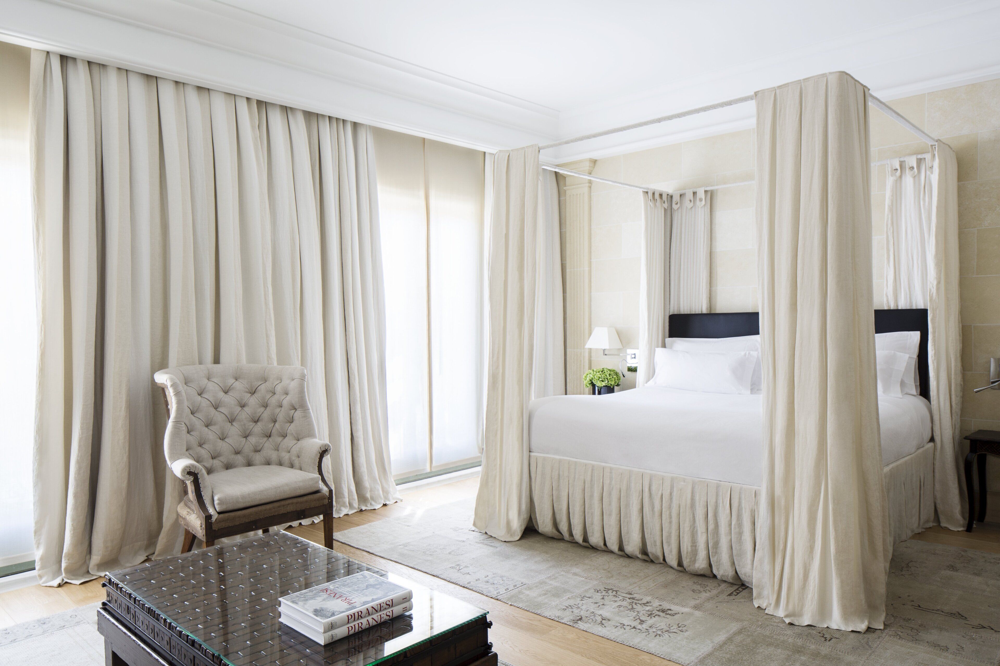 Una cama con dosel y cuatro columnas, un sillón capitonado, una mesita de noche con libros y un ventanal con cortinas.