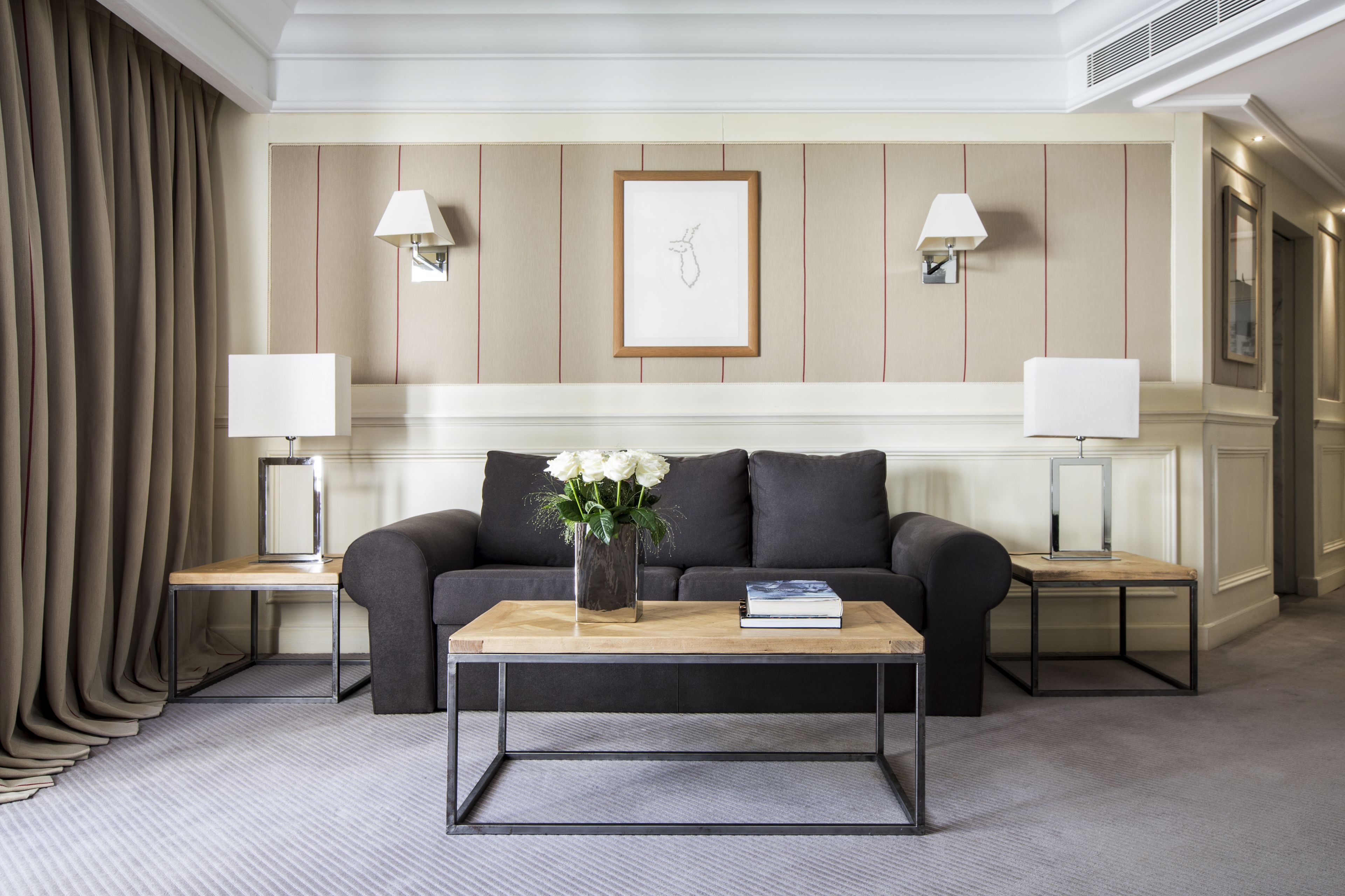 A modern living room with a dark grey sofa, a wooden coffee table, and two bedside tables with lamps. A framed picture hangs on the wall above the sofa.