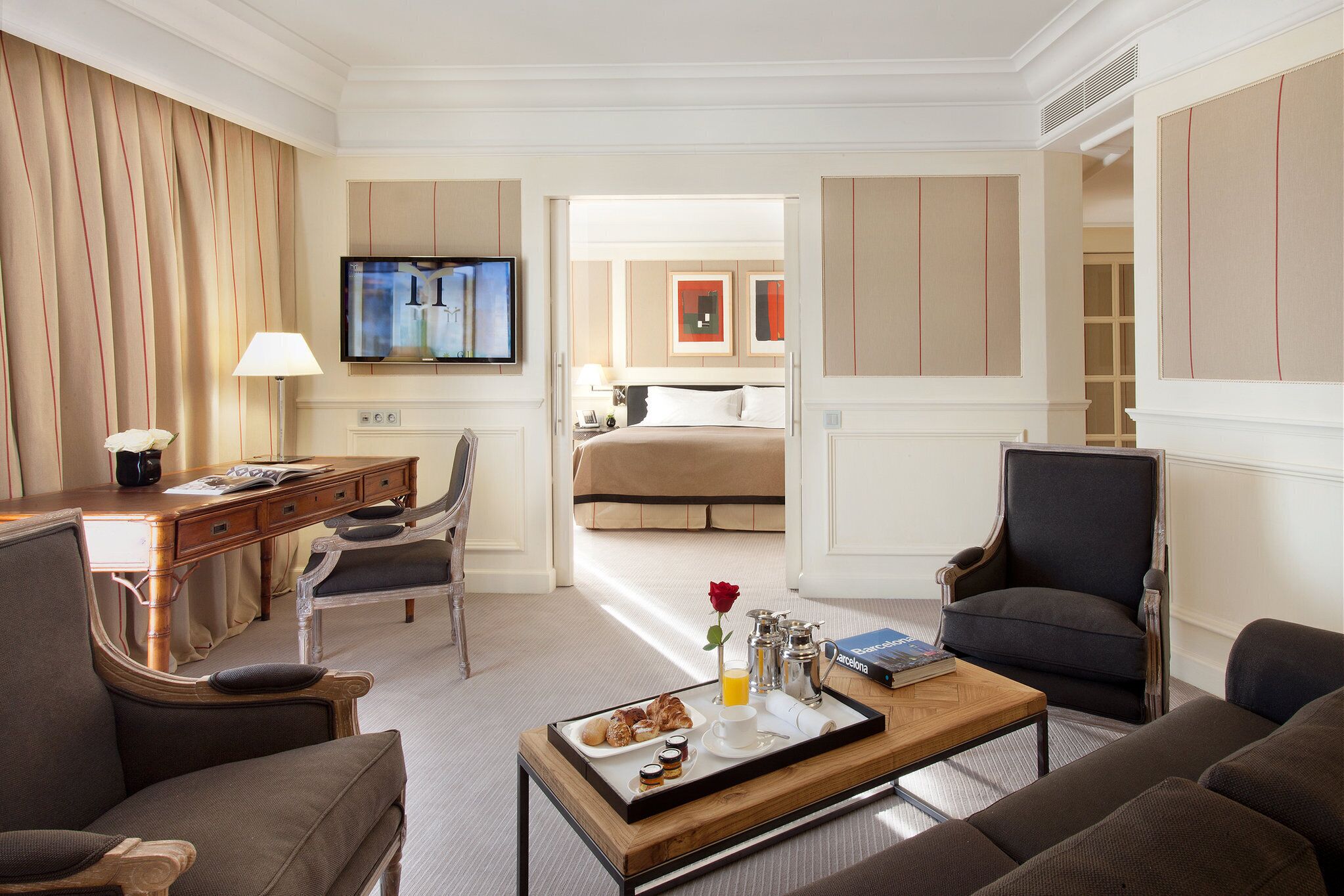 A hotel room with a bed, a desk with a lamp, a coffee table with breakfast items, and a TV on the wall.